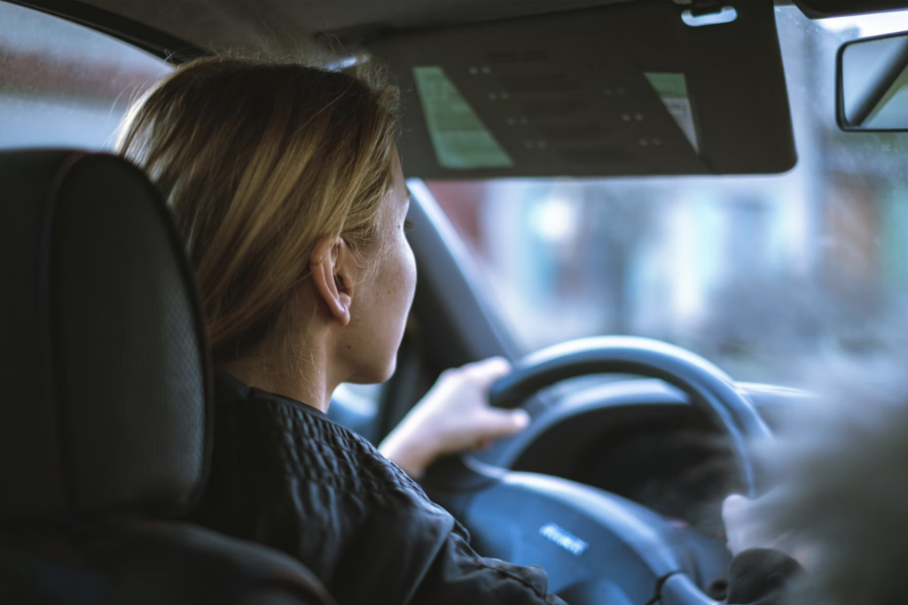 A woman with blonde hair drives a car, focused intently on the road. The interior is slightly blurred, suggesting motion. The scene feels calm and steady.