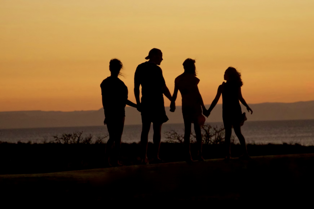 Silhouetted family of four holding hands, standing together at sunset by the sea. The sky glows orange, creating a warm, serene atmosphere.